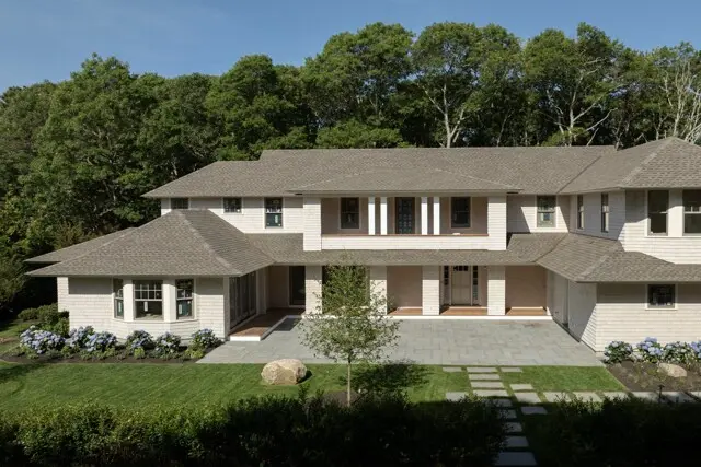Two-story light-brick house with a gray roof, centered entry, manicured lawn, stone walkway, and hydrangeas along the front, with trees in the background.