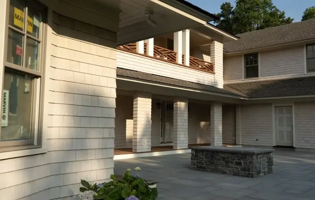 Two-story house with light siding, a covered porch on columns, a wooden balcony, and a stone fire pit in a paved courtyard; hydrangeas in the foreground.