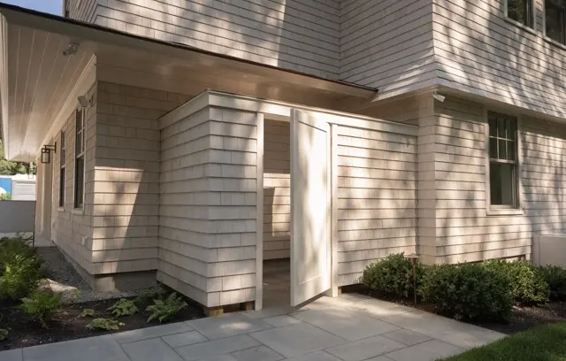 Light beige clapboard house with an open entry door, side window, and landscaped front shrubs; a paved walkway under dappled sunlight.