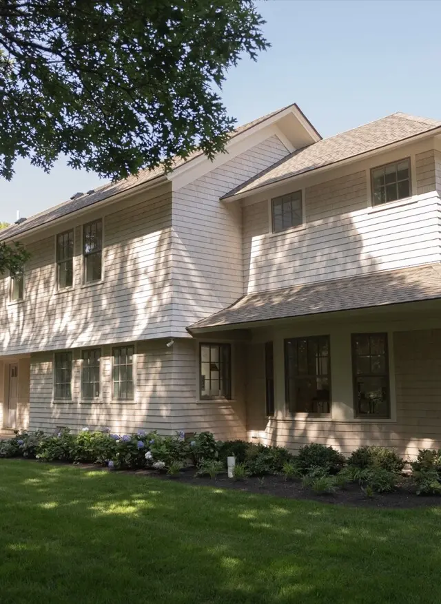 Two-story beige house with light siding, many windows, a front porch, and a manicured lawn with shrubs along the foundation, shaded by trees.
