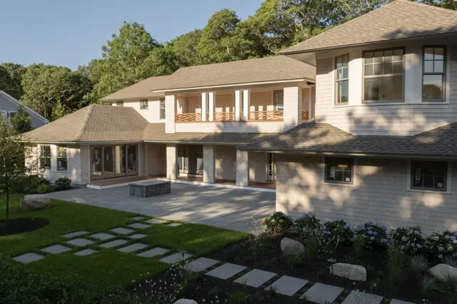 Two-story beige brick house with a tiled roof, large windows, and a second-floor balcony; a patio with a stone fire pit and stepping-stone path.