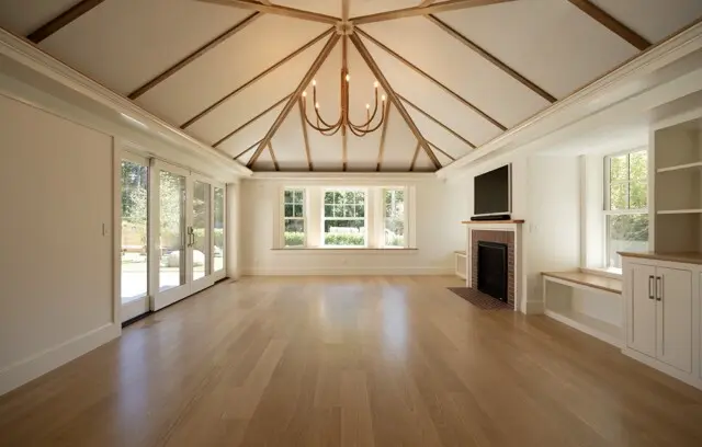 Bright living room with vaulted ceiling and exposed wood beams, central chandelier, large glass doors, window seat, fireplace with TV, and built-in cabinets.
