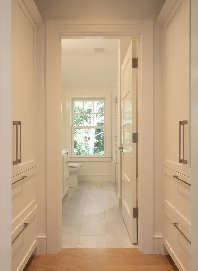 Bright white hallway with built-in cabinets on both sides opening to a sunlit bathroom with a window and toilet.