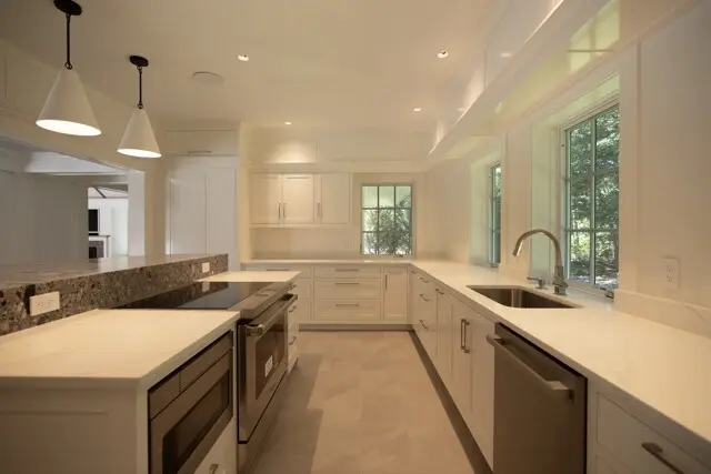 Bright white kitchen with marble countertops, stainless appliances, an island with a stove, pendant lights, and a sink by windows overlooking greenery.