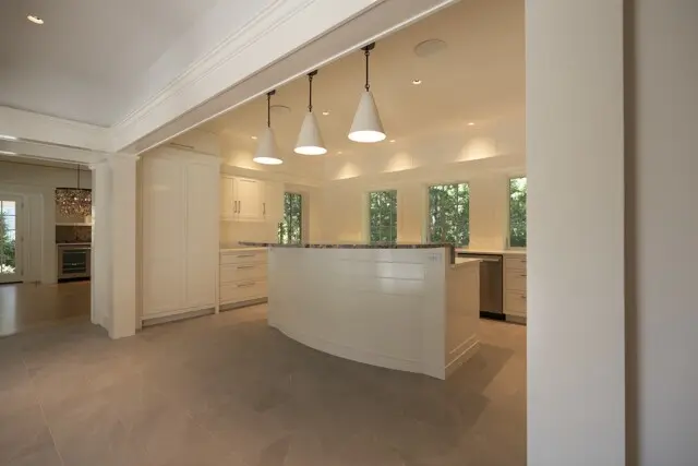 Bright white kitchen with curved island, white cabinets, three pendant lights, large windows, stainless appliances, and light gray tile floor.