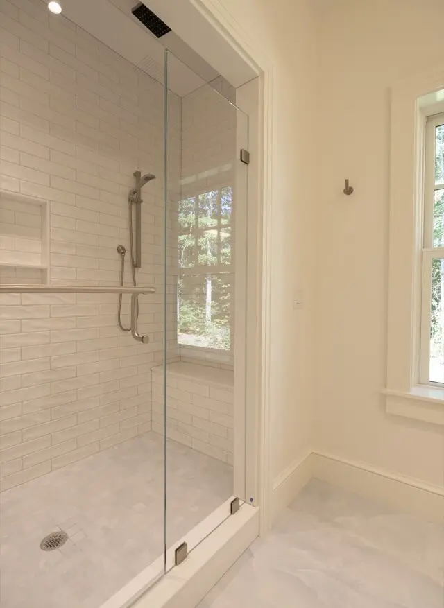 Modern glass shower with white subway tiles, rainfall and handheld showerheads, built-in niche, and a window showing trees outside in a bright bathroom.
