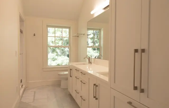 Bright white bathroom with a double-sink vanity, sleek hardware, tall cabinets, and a large mirror. A window over the toilet frames greenery outside.