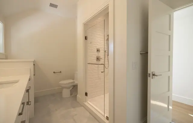 Bright, modern bathroom with a white vanity on the left, glass-enclosed shower, and a toilet against the back wall; light gray tile floor.