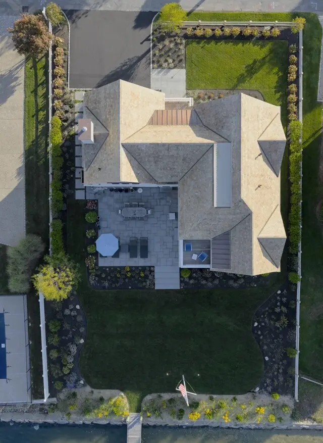Aerial view of a beige-roofed house with a large curved lawn, back patio with furniture, a flagpole with American flag, and a dock reaching the water.