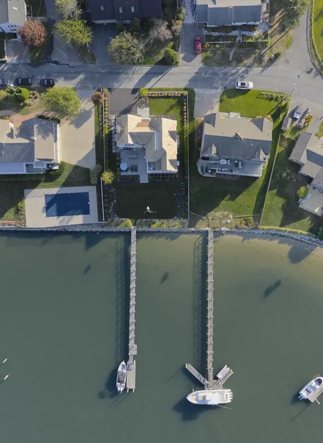 Aerial view of a waterfront neighborhood: houses along a street with green lawns, and two long piers extending into calm water with boats moored at their ends.