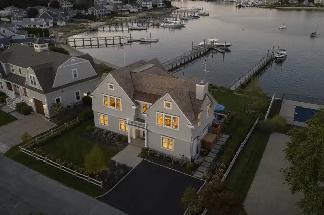 Aerial view of a coastal neighborhood with a light-gray two-story house and gabled roof beside a marina, showing docks, boats, and calm water.