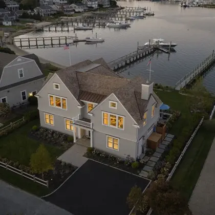 Aerial view of a coastal neighborhood with a light-gray two-story house and gabled roof beside a marina, showing docks, boats, and calm water.