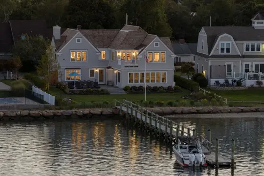 Evening view of a gray two-story waterfront home with warm interior lights, a wooden dock reaching a motorboat, and water reflecting the glowing windows.