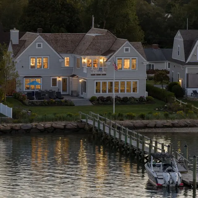 Evening view of a gray two-story waterfront home with warm interior lights, a wooden dock reaching a motorboat, and water reflecting the glowing windows.