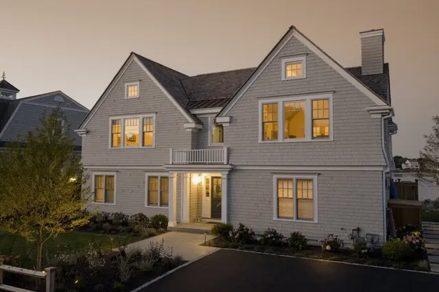 Two-story gray house with steep gables, warm lit windows, and a front porch light; landscaped yard with a tree left and a paved driveway at dusk.