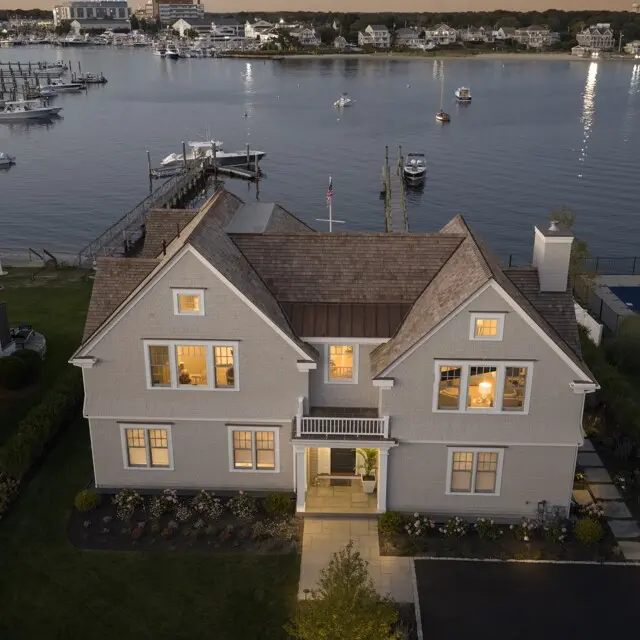 Two-story gray coastal home with peaked roofs sits by a marina; boats dot the water and warm lights glow from the windows.