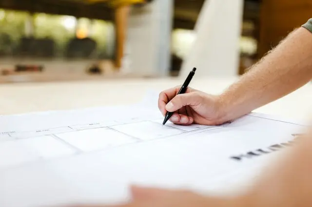 Close-up of a hand holding a black pen and drawing on architectural blueprints spread on a table, with a blurred indoor background.