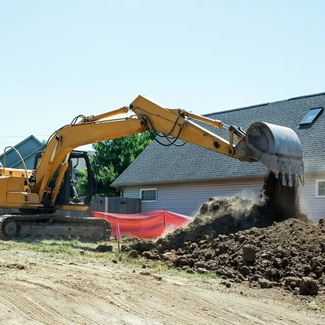 Yellow excavator with extended arm dumping soil onto a mound at a construction site beside a suburban home, with orange safety fencing nearby.