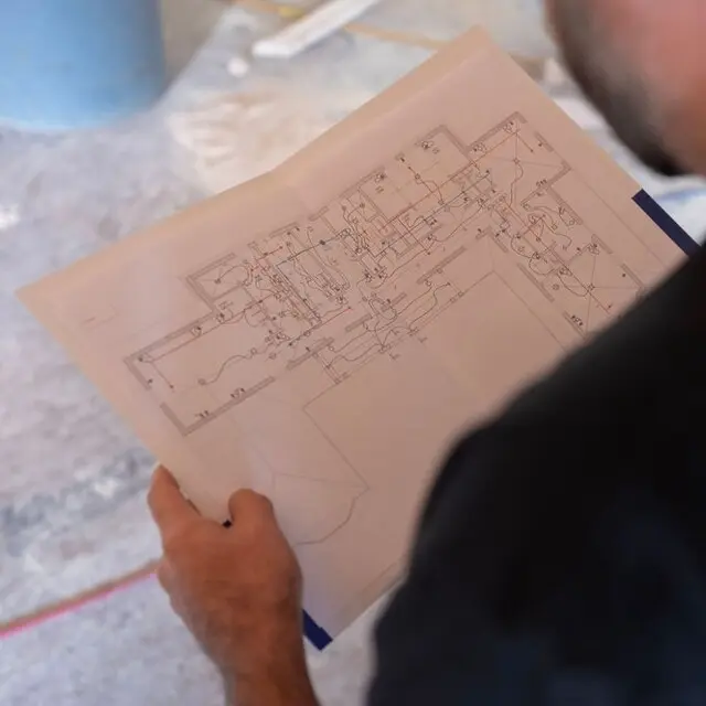 A person holds a printed architectural blueprint with red and blue lines, inspecting a building plan on a dusty concrete floor indoors.