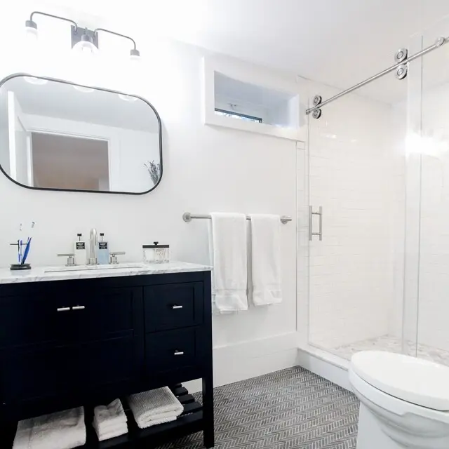 Modern bathroom with a dark vanity and marble countertop, rounded mirror, towel rack with white towels, glass shower enclosure, and a white toilet.