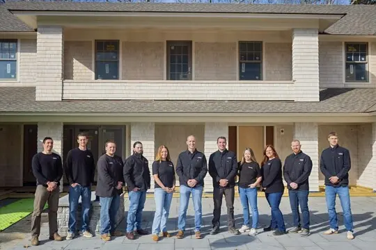 Line of construction crew members in black shirts stand in front of a two-story house with a white-brick exterior and a new porch under construction.