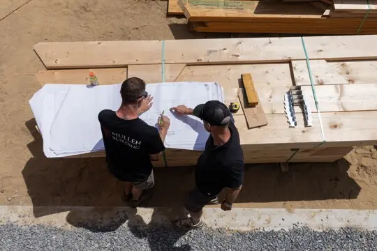 Top-down view of two workers examining blueprints on a white sheet over a lumber stack at a construction site; tools nearby.