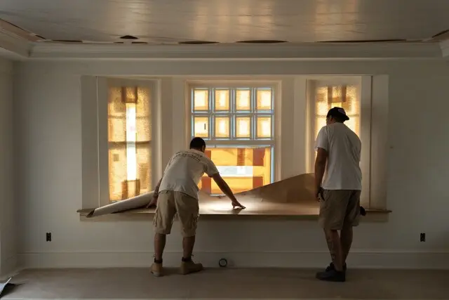 Two people in light shirts and shorts unroll brown wallpaper over a window ledge in a sunlit, renovated room; orange-tinted window panes glow behind them.
