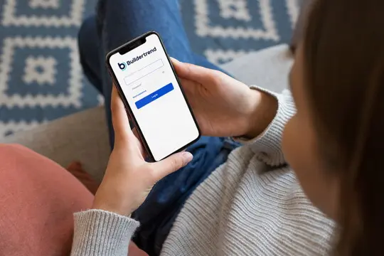 Person sits on a couch, holding a smartphone displaying a Buildertrend login screen with fields for email and password and a blue Log In button.