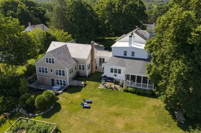 Aerial view of a sunny residential yard with two large houses, green lawn, trees, outdoor seating, and a screened porch.