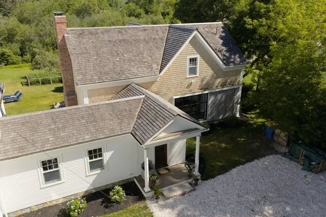 Aerial view of a two-story beige and white house with a steep roof, brick chimney, front porch, gravel driveway, and surrounding green yard and trees.