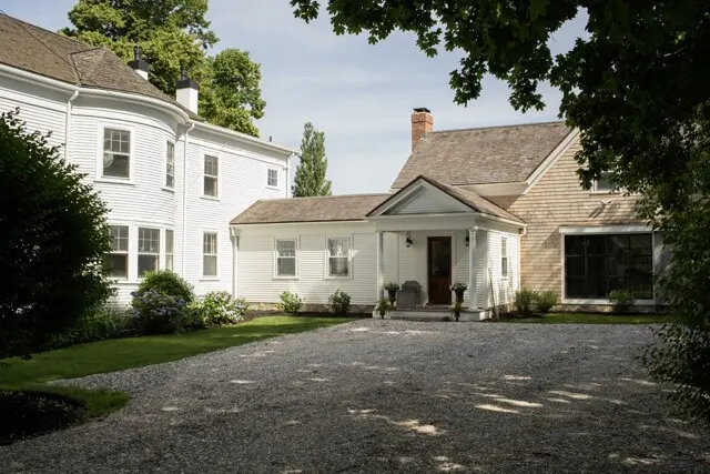 White, two-story house with a front porch and gabled roof, set beside a gravel driveway and greenery; combines white siding with a brick-addition on the right.
