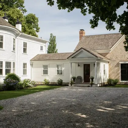 White, two-story house with a front porch and gabled roof, set beside a gravel driveway and greenery; combines white siding with a brick-addition on the right.