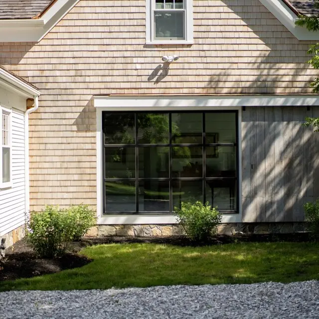 Front view of a beige shingle house with a large grid window, white trim, and a small covered porch on the left; greenery and gravel path in front.