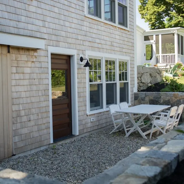 A light wooden house with a dark brown door, large windows, and an outdoor white table with matching chairs on a gravel patio; a stone wall and garden in the background.