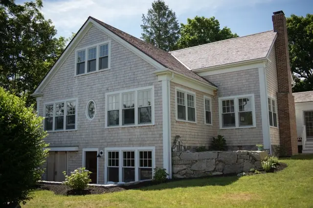 A light wooden house with multiple white-framed windows, a gabled roof, circular window, stone foundation, and a brick chimney amid green trees and lawn.