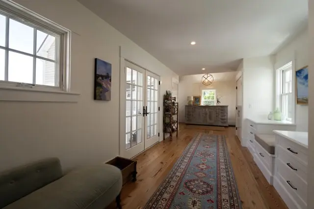 Bright hallway with light wood floor, white walls, glass French doors, built-ins, a patterned runner rug, and a gray dresser at the far end; natural light through windows.