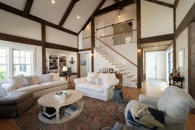 Bright living room with white sofas, wooden beams, staircase, and large windows; cozy seating on a patterned rug with a round coffee table and books.