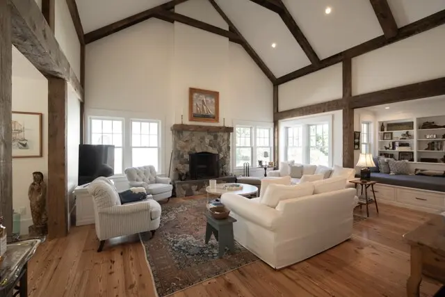 Bright living room with white sofas, a stone fireplace, wooden beam ceiling, large windows, and a built-in window seat. Cozy, open family area.