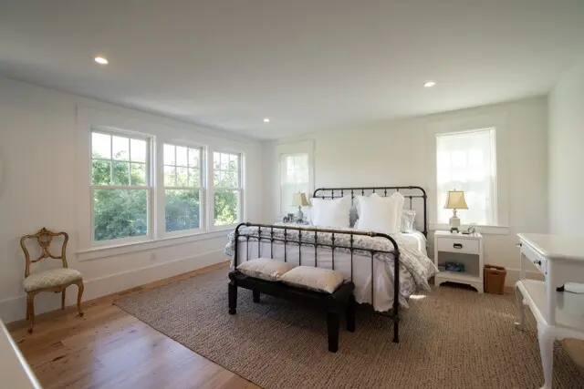Bright, airy bedroom with a vintage metal bed, white bedding, two nightstands, lamps, and a woven rug; large windows with greenery outside.