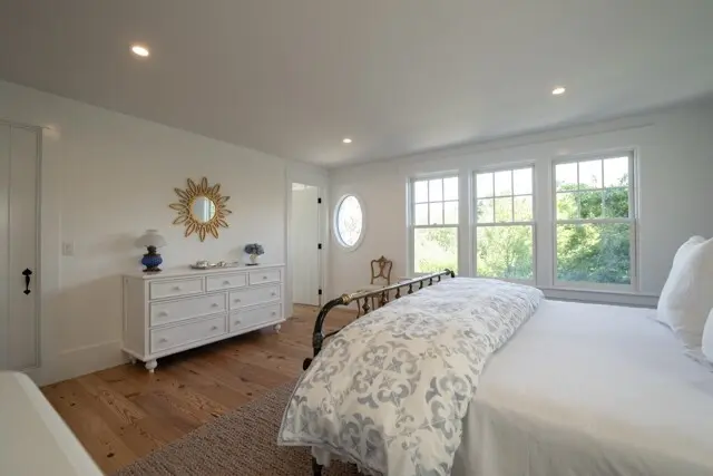 Bright master bedroom with a white dresser, sunburst mirror, and a round window. A wrought-iron bed with a pale blue patterned comforter faces large windows overlooking greenery.