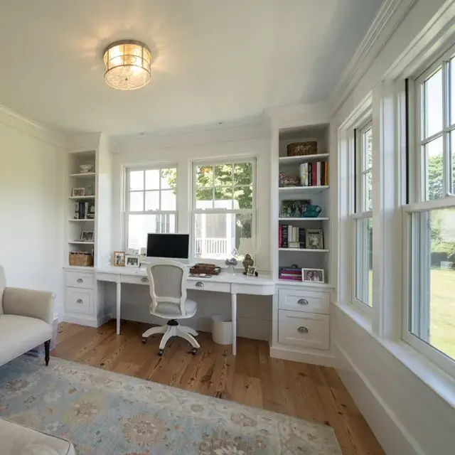 Bright home office with white built-in shelves, desk, and chair by large windows; beige sofa, light rug, and hardwood floors. Sunny, organized space.