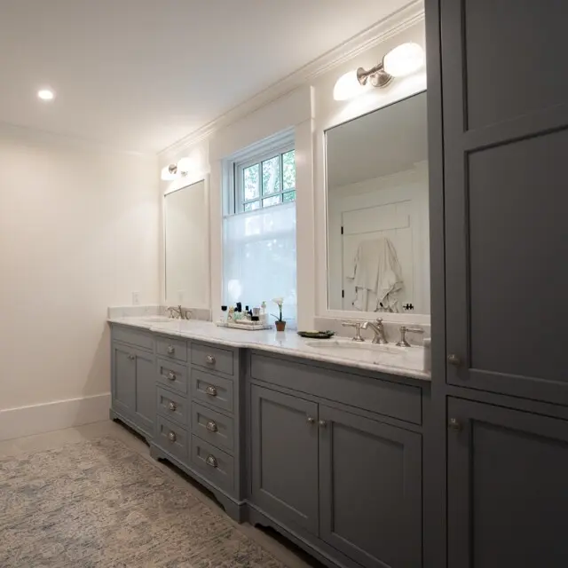 Bright, airy bathroom with light gray cabinetry, marble countertops, double sinks, and two mirrors. A window brings in natural light; a rug covers tiled floor.