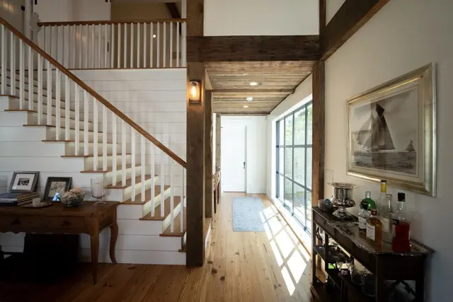 Bright foyer with a wooden staircase on left, white railing, and open hallway. Sunlight streams through large windows on right, bar cart and framed art on wall.
