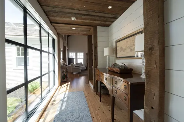 Bright farmhouse hallway with large windows on left, rustic wooden beams, and a curving wooden console table with lamps and a framed map.