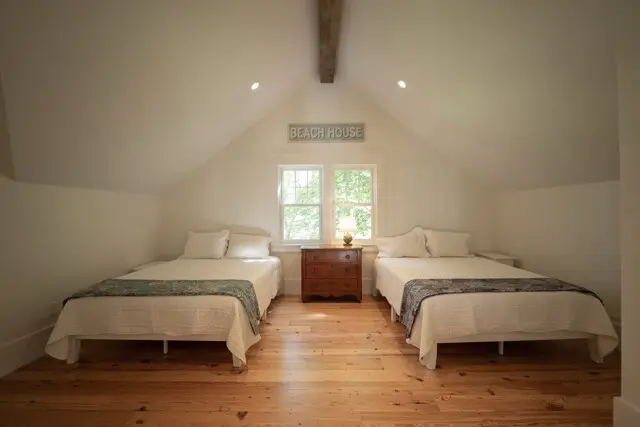 Sunlit attic bedroom with two twin beds, white quilts and teal runners, a wooden dresser between windows, “BEACH HOUSE” sign above, and a small lamp on top.