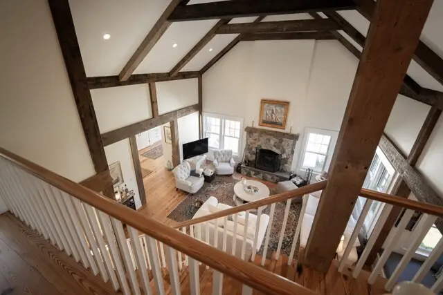 View of a bright living room from a mezzanine: exposed wooden beams, stone fireplace, white seating, large windows, and a rug-centered coffee area.