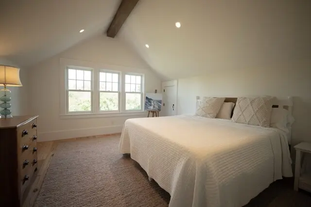 Bright, cozy attic bedroom with a white quilted bed, two cream textured pillows, wooden dresser, and a lamp; three-pane window with greenery outside. Ceiling beam.