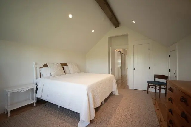 Bright airy bedroom with a white bed, matching pillows, and a white side table. Sloped ceiling, exposed wooden beam, and light wood floor. Dresser and chair in corner.