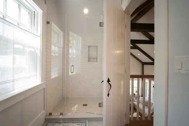 Bright white tiled bathroom with a glass-enclosed shower, chrome hardware, and built-in niche. Sloped ceiling with dark wooden beams visible beyond.