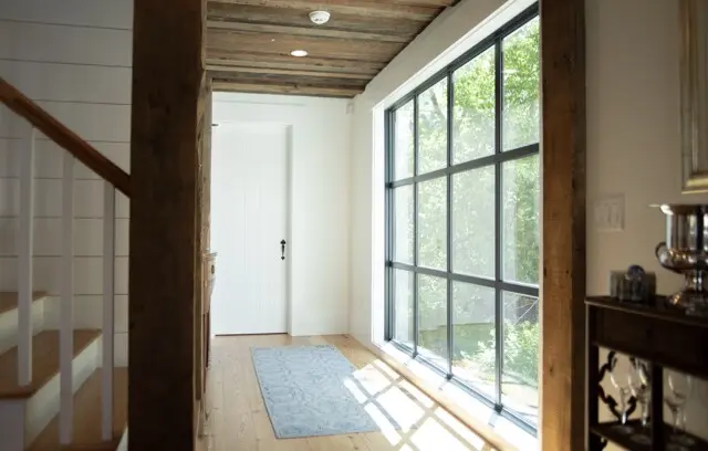 Bright hallway with large grid windows on right, white door at end, rustic wooden beams and stairs on left, light wood floor, pale blue rug.